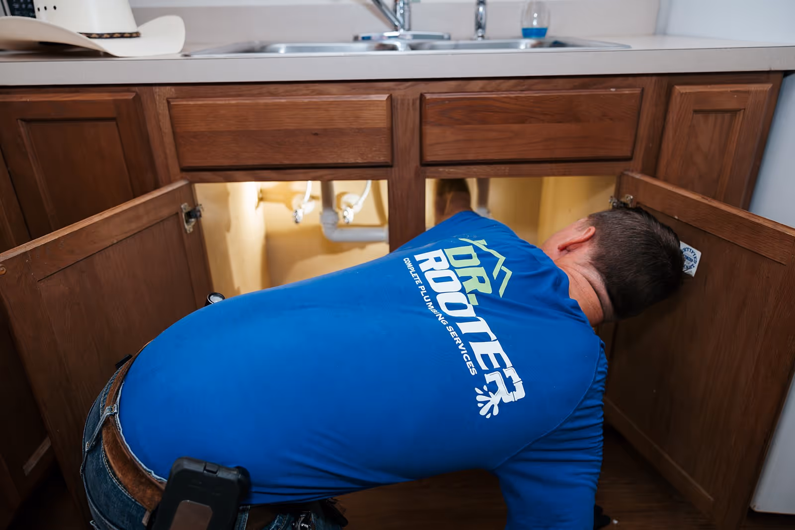 A plumbing wearing a blue uniform kneels by a kitchen sink, using his tools to unclog the drain.