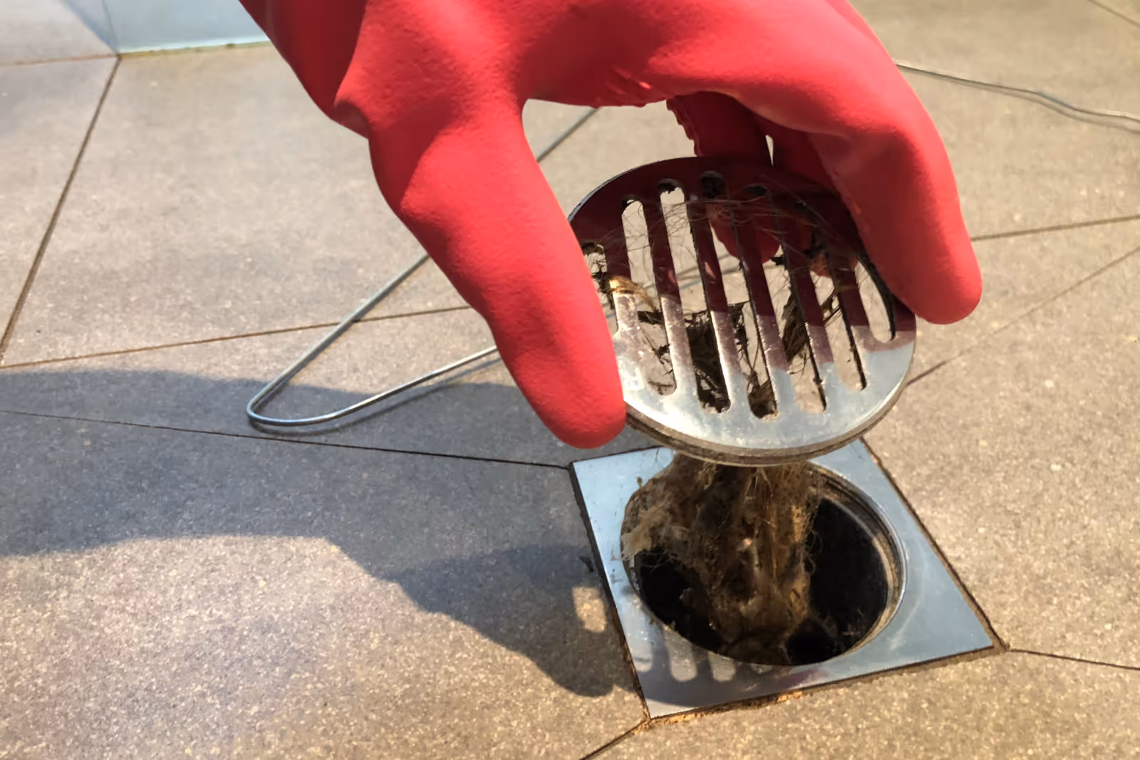 A plumber in red gloves pulls off a drain cover to reveal a clogged drain underneath.