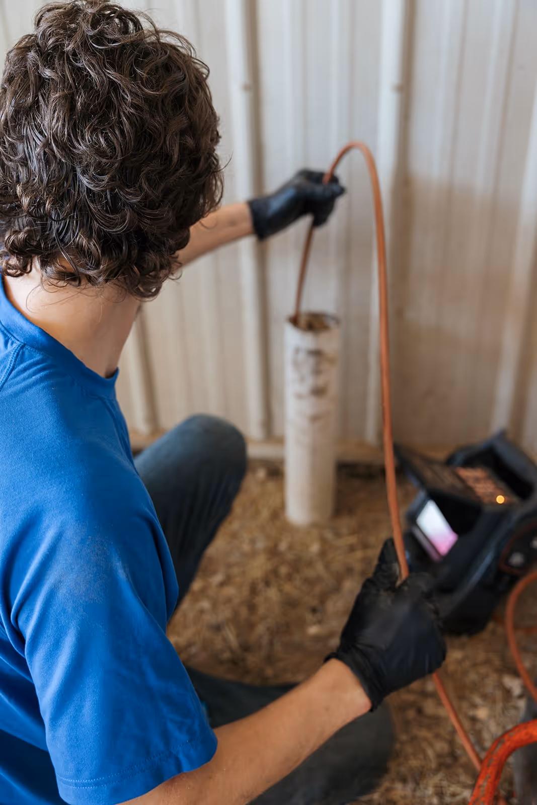 A plumber crouches under a sink to fix a leaking pipe. The plumber wears a blue uniform and is focused on the task at hand.