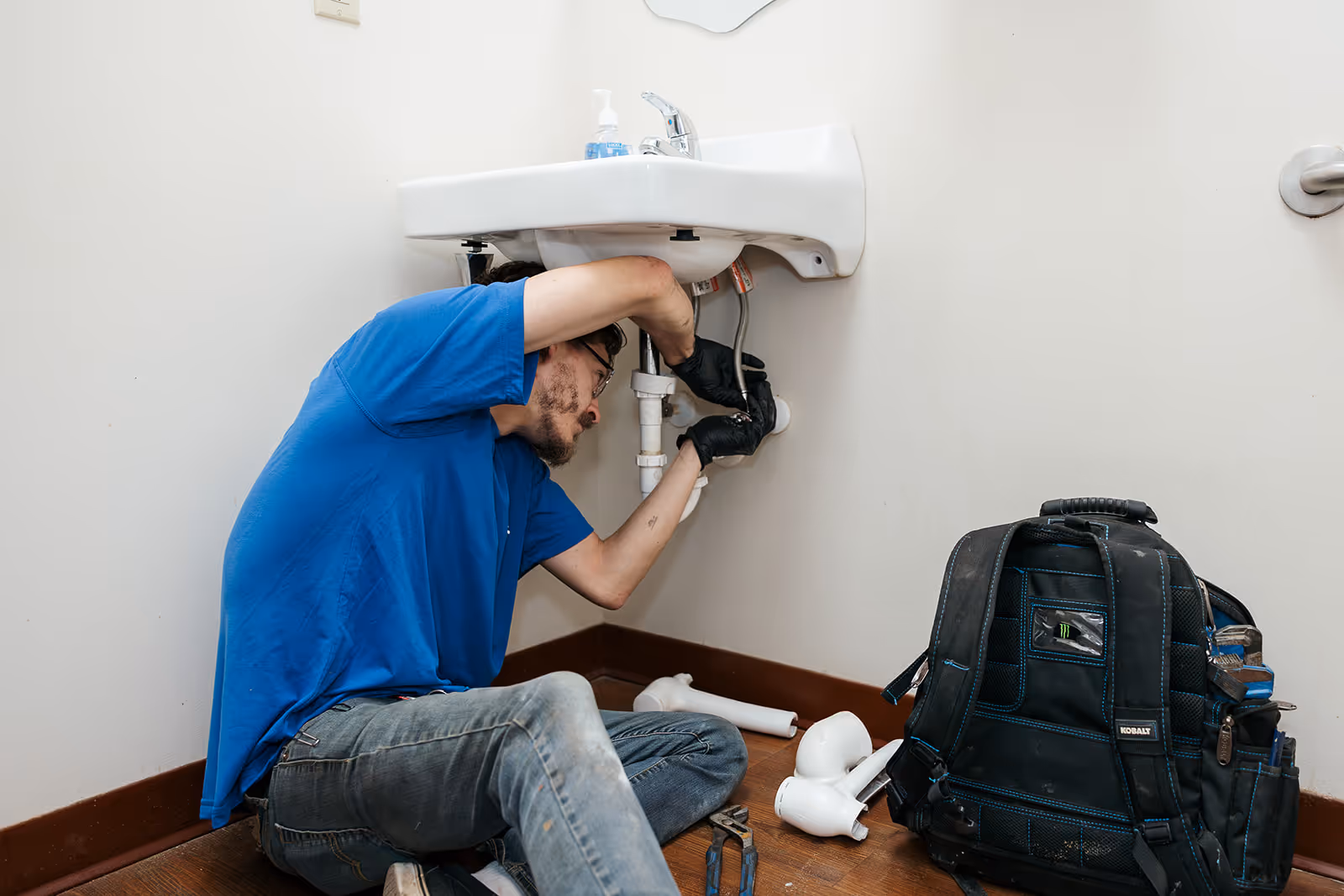A plumber wearing a blue top inspects a drain, with a black bag and his tools placed on the ground beside him.